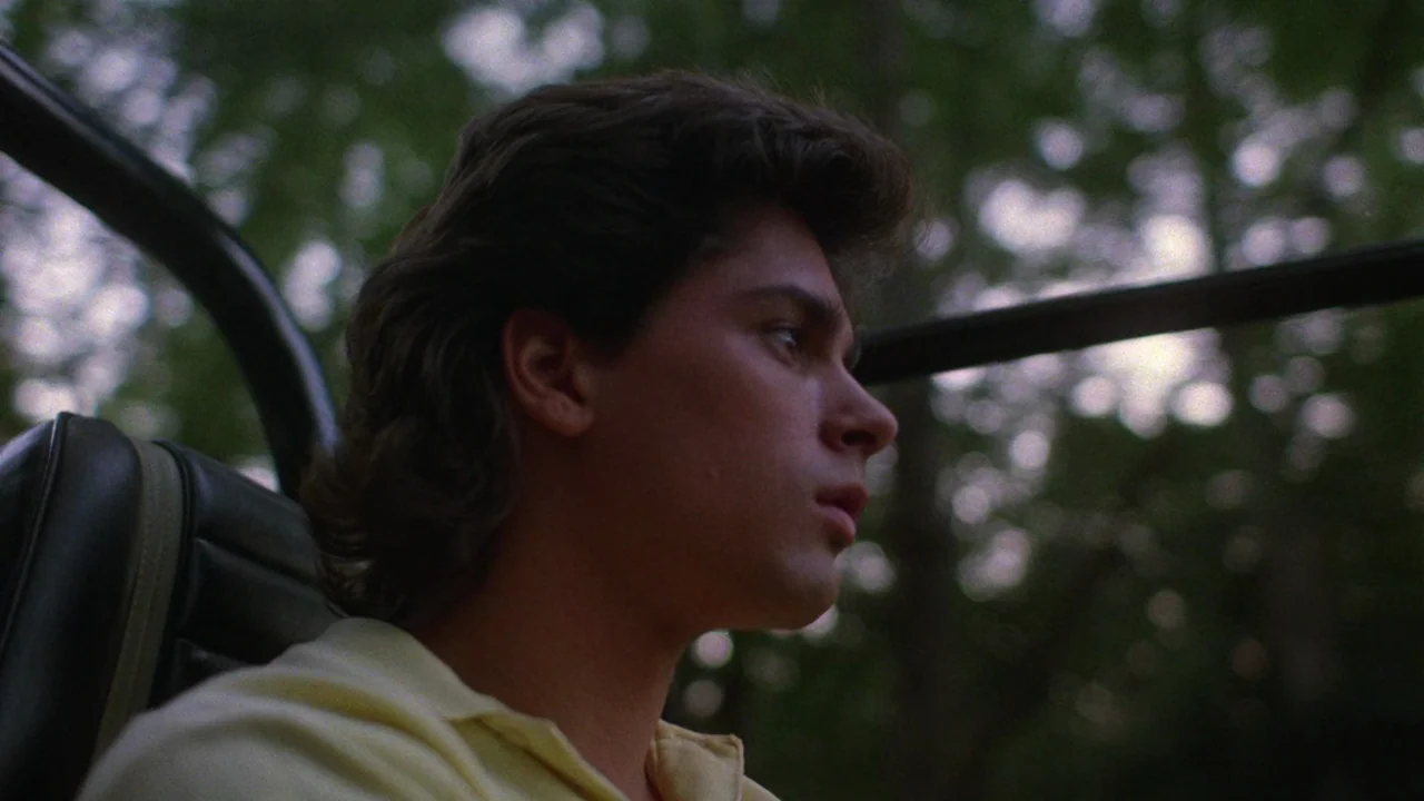Society (1989) movie still: closeup — The interior of a convertible car features a black leather headrest and metal frame. A young man sits in the…
