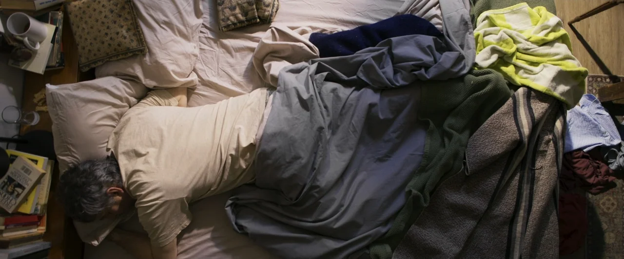 Dog Life (2024) movie still: medium — A top-down view of a cluttered bed with layered blankets and scattered books. A middle-aged man lies asleep on…