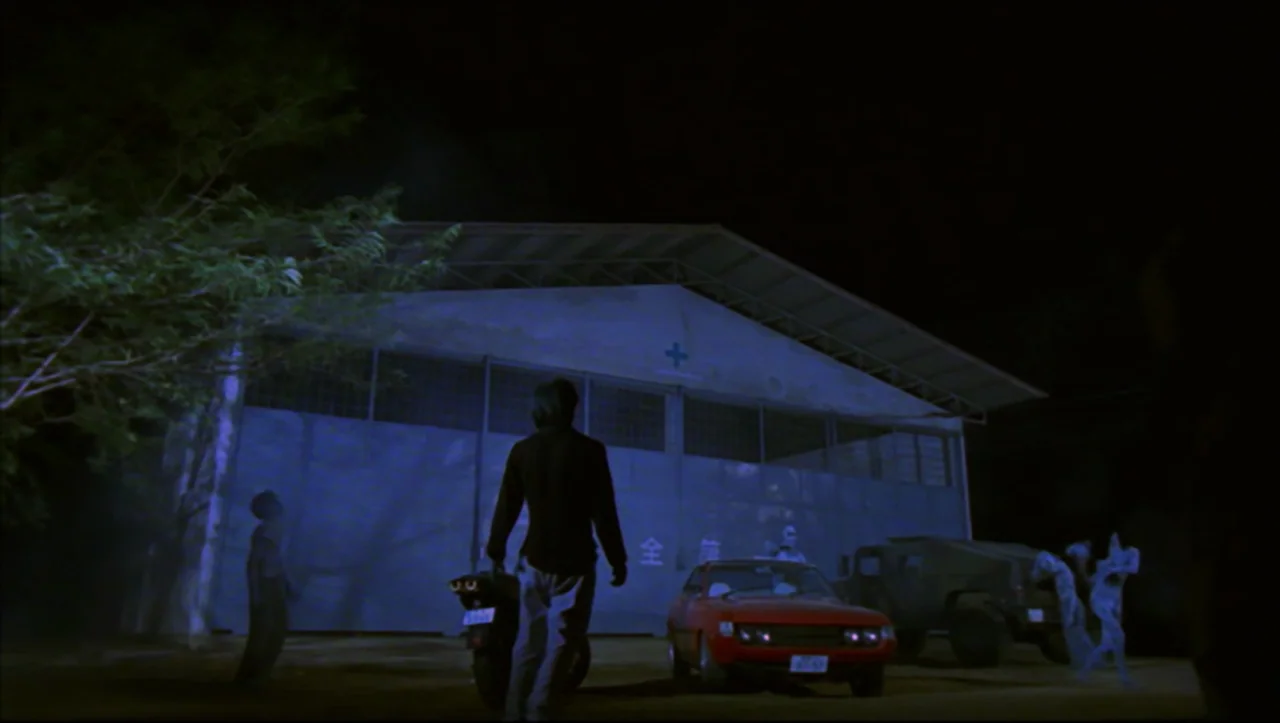 Wild Zero (1999) movie still: wide — A large industrial building with high metal doors stands at night under dark skies. A young man walks away from…