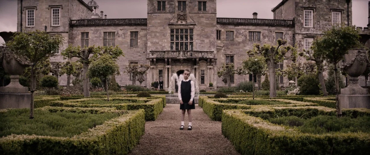 Tomb Raider (2018) movie still: fullbody — A stone manor house stands behind a symmetrical garden with manicured boxwood hedges. A young girl stands…