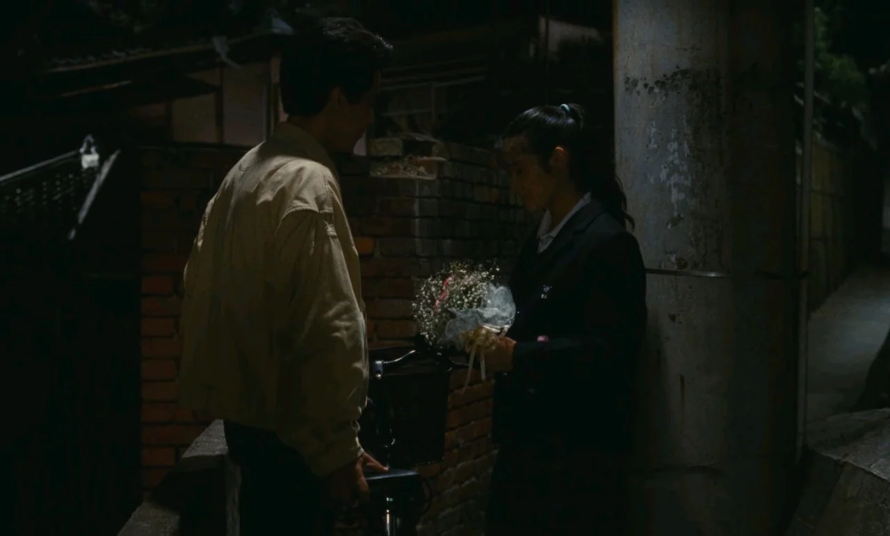Chizuko’s Younger Sister (1991) movie still: medium — A dimly lit narrow alleyway features a red brick wall and concrete pillars. A young couple…