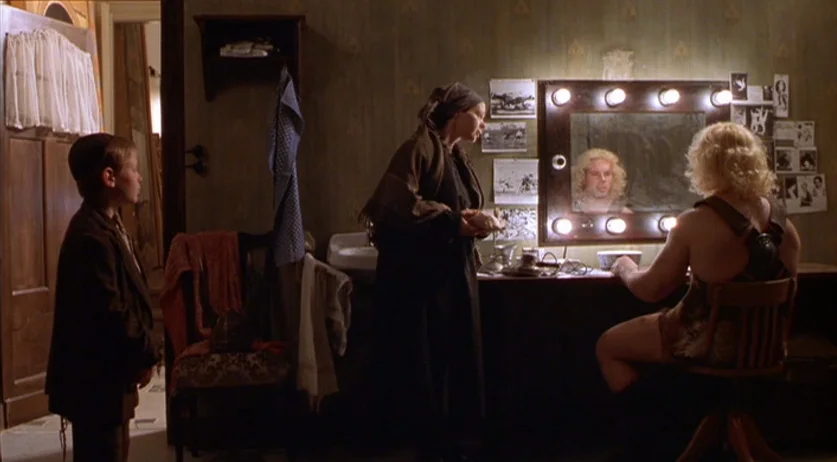 Invincible (2001) movie still: wide — A dimly lit backstage dressing room features wallpaper, vanity photos, and an illuminated mirror. A woman in…