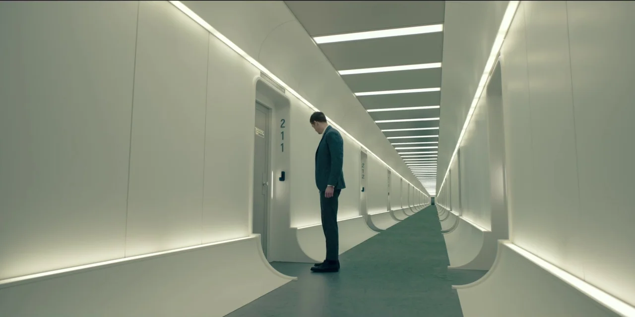 Mute (2018) movie still: fullbody — A long, minimalist corridor with white curved walls and a receding perspective point. A man in a dark suit…