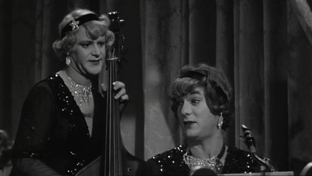 Some Like It Hot (1959) movie still: medium — Two men in 1920s-style evening dresses and wigs perform on a stage with curtains. They are positioned…