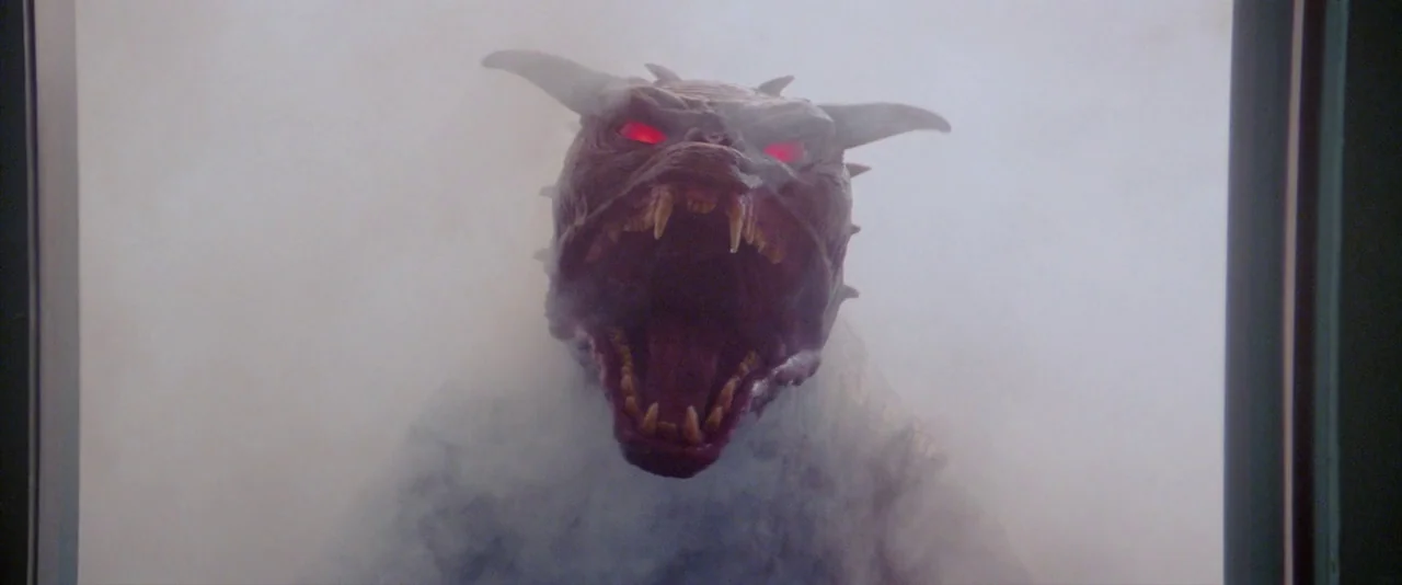 Ghostbusters (1984) movie still: closeup — A dark, fog-filled space is viewed through a glass pane. A monstrous creature with glowing red eyes…