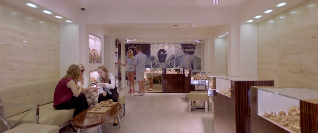 Holiday movie still: wide — An interior shot of a modern luxury jewelry store with beige marble walls. Three women sit on a leather sofa examining…