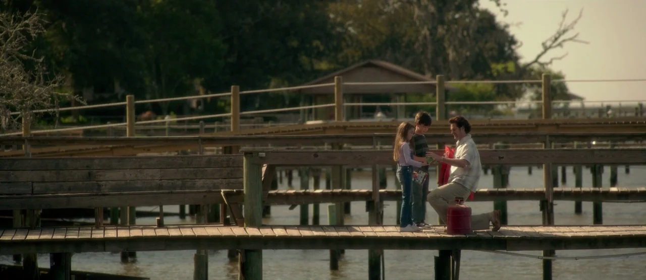 Gerald’s Game (2017) movie still: wide — A wooden pier extends over calm water with a simple covered structure in the distance. An adult man kneels…