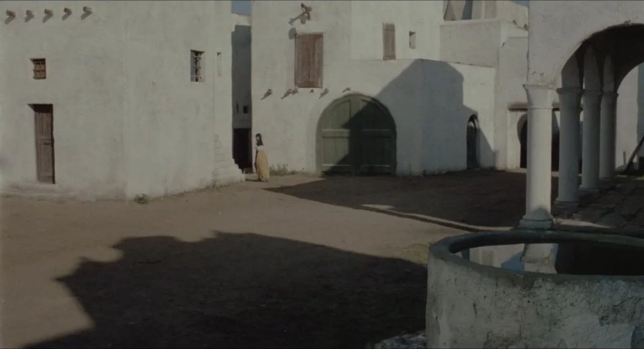 A Stranger in Town (1943) movie still: wide — A Mediterranean village square features white plastered buildings with brown wooden doors and columns.…