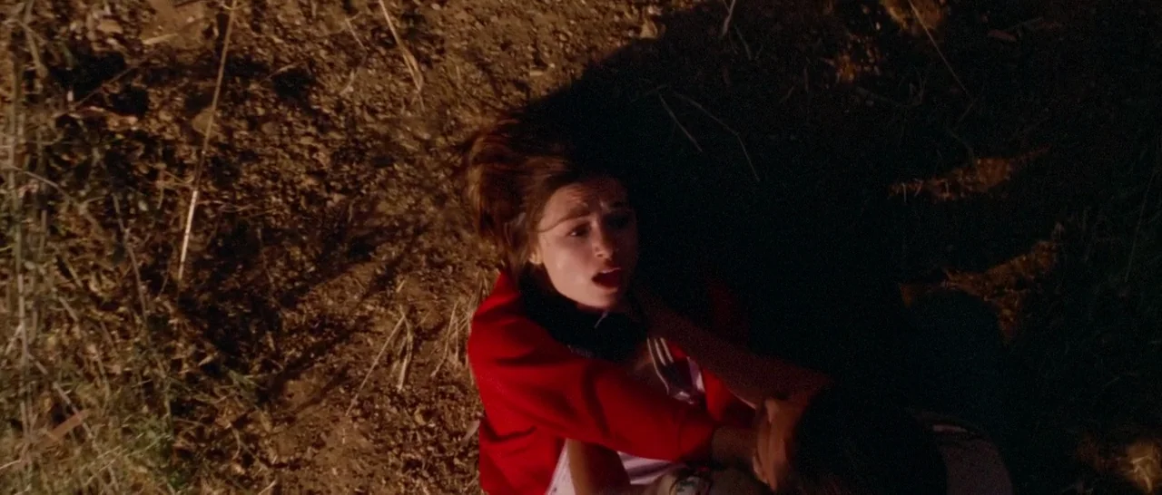 Too Late (2015) movie still: medium — A woman lies on dark dirt with patches of dry grass and debris. A person stands over her gripping her neck…