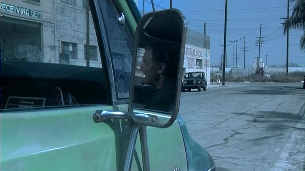 Land of Plenty (2004) movie still: medium — An industrial street is visible with a warehouse and utility poles under a bright sky. A man sits inside…