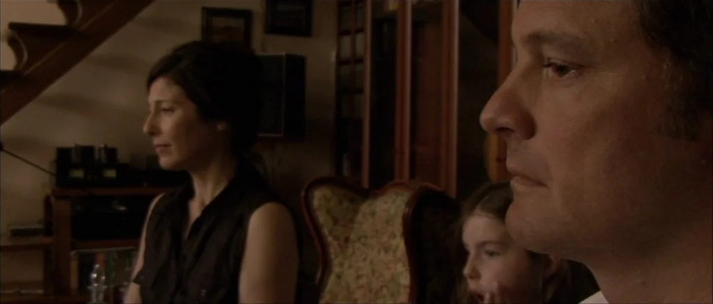 Genova (2008) movie still: medium — A dimly lit room features a wooden staircase and a bookshelf in the background. A man and a woman sit together…