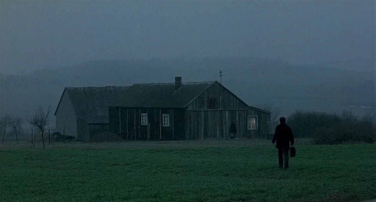 Three Colours: White (1994) movie still: wide — A dark wooden farmhouse sits in a misty, flat field during blue hour. A man walks across the…