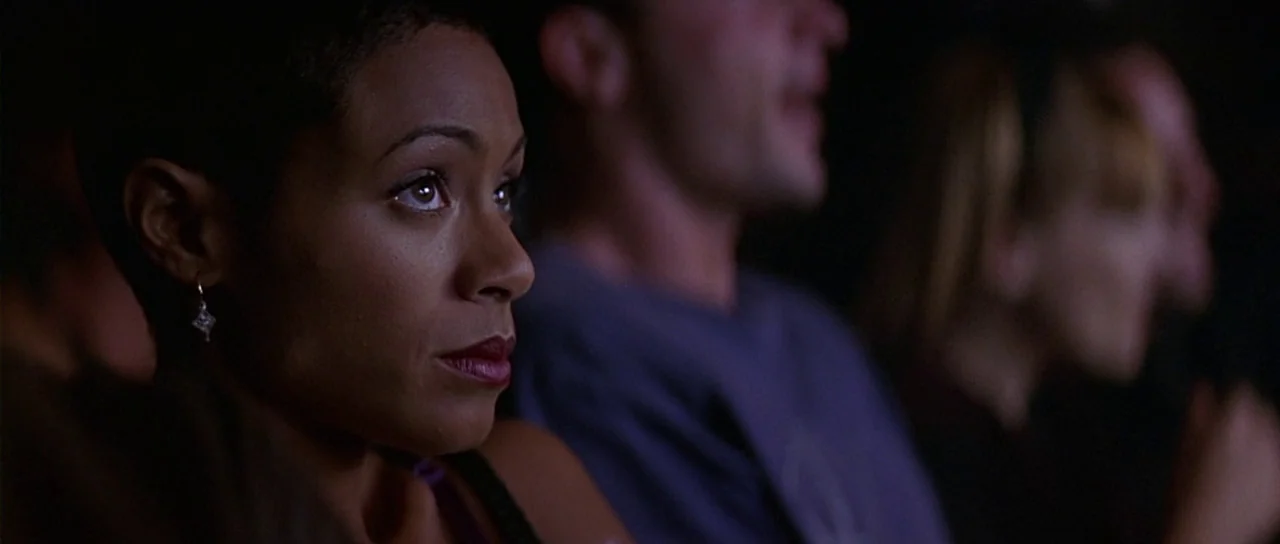 Scream 2 (1997) movie still: closeup — A dimly lit movie theater auditorium with audience members seated in rows. A young woman with short hair…