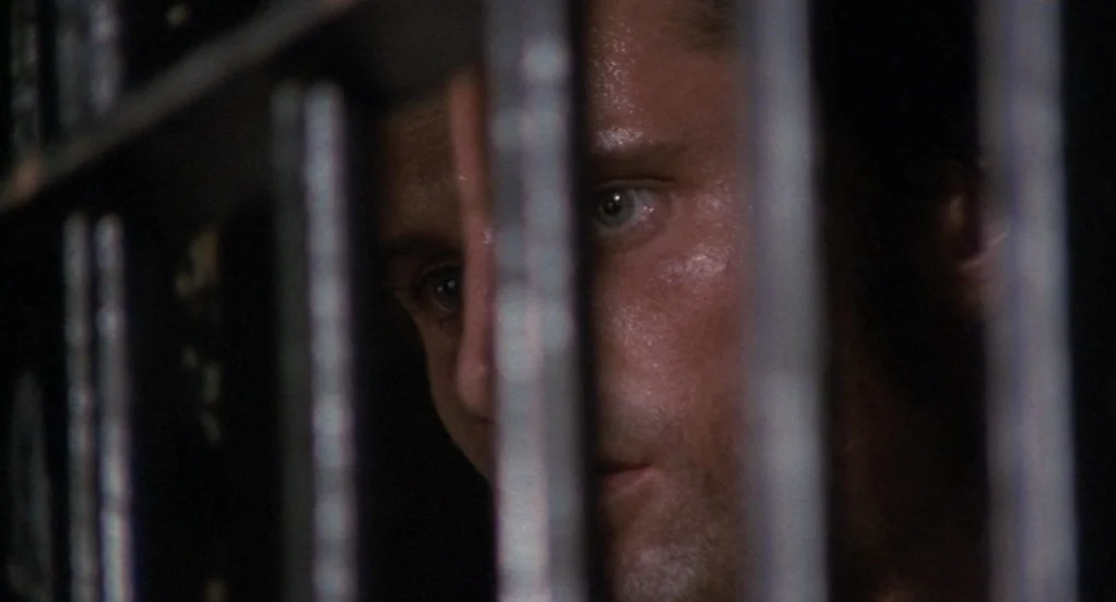 The Indian Runner (1991) movie still: closeup — A man is viewed through the dark vertical bars of a metal holding cell. The subject watches through…