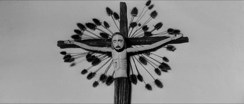 Marketa Lazarová (1967) movie still: closeup — A wooden crucifix is mounted against a plain white wall. The cross features dried thistle-like seed…