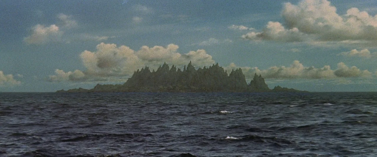 King Kong Vs. Godzilla (1962) movie still: establishing — A dark, craggy island rises out of a vast expanse of dark blue water. The island has…