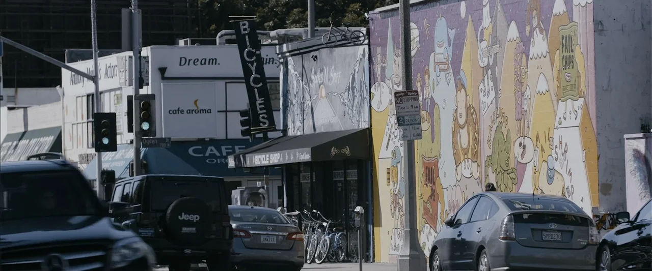The Kleptocrats (2018) movie still: wide — A city street in daytime features a bicycle shop and a large painted mural. Several cars and a Jeep are…