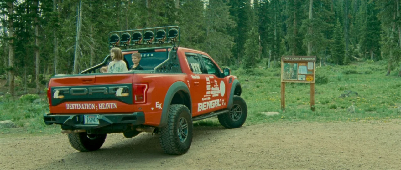Riddle of Fire (2023) movie still: wide — A bright orange pickup truck is parked on a dirt path beside a forest. Two children sit in the truck bed…