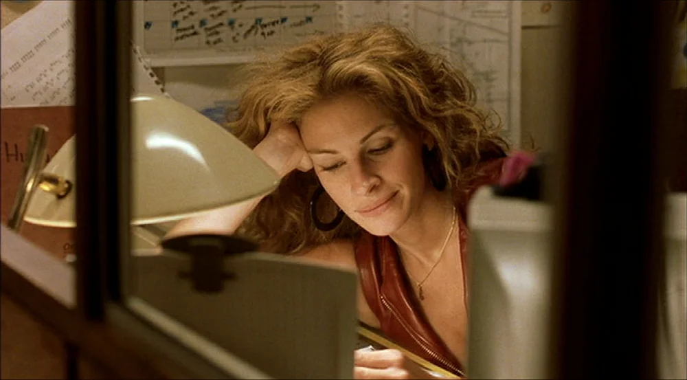 Erin Brockovich (2000) movie still: closeup — A workspace featuring a desk lamp and paper documents pinned to a partition. A woman sits at her desk…