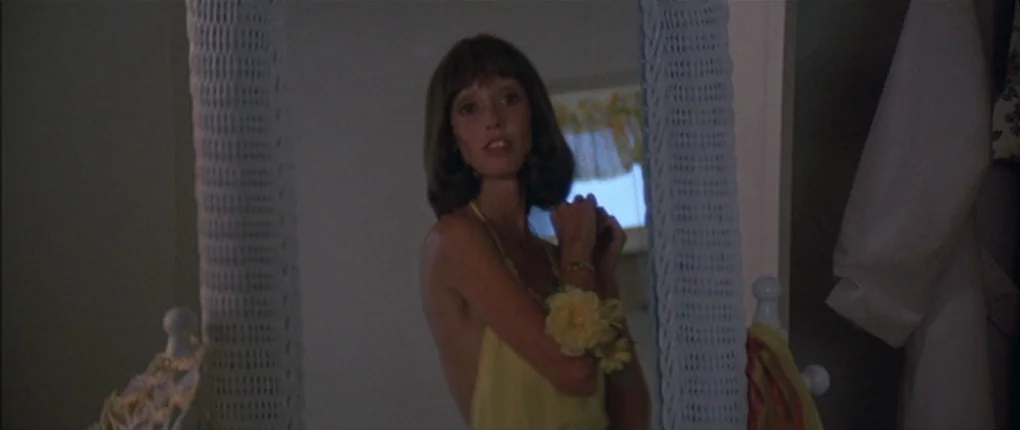 3 Women (1977) movie still: medium — A woman stands before a vanity mirror with a textured blue-white border. She adjusts a floral corsage on her…