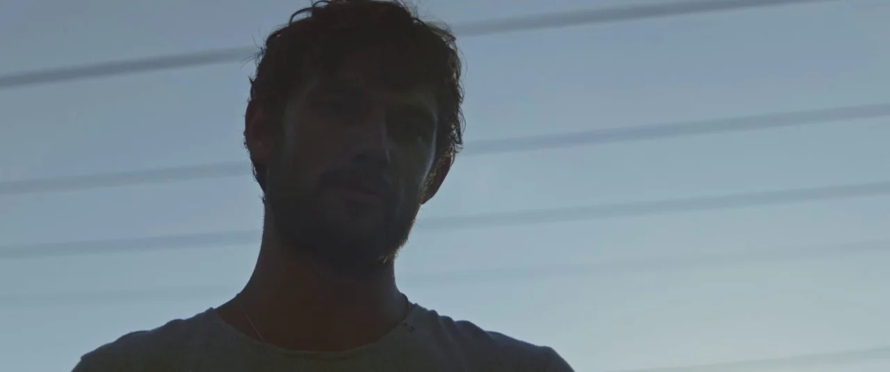 The Strange Ones (2017) movie still: closeup — A man stands outdoors at dusk against a clear, pale blue sky background. The subject is backlit with…
