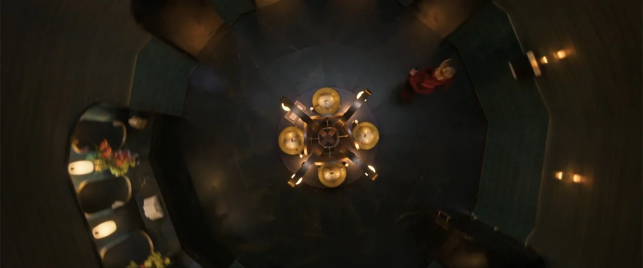 Drop (2025) movie still: establishing — A modern, dimly lit public restroom features dark tiled walls and a central circular sink unit. The space is…