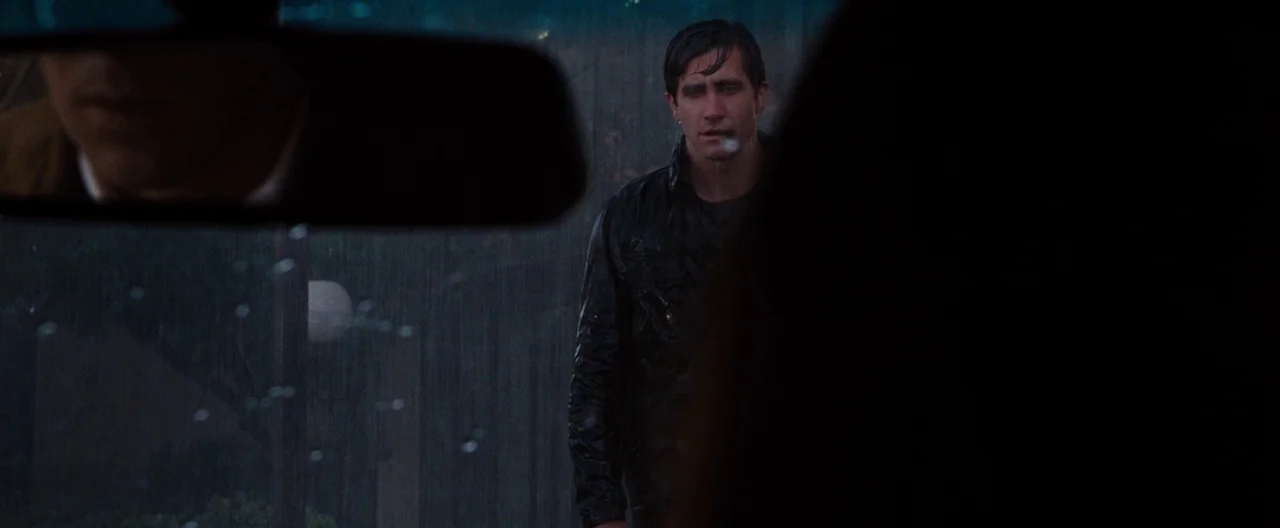 Nocturnal Animals (2016) movie still: medium — Rain falls steadily outside a vehicle at night in a dimly lit residential area. A man stands in the…