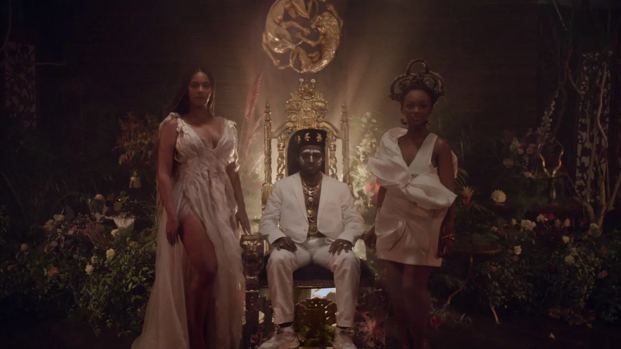 Black is King (2020) movie still: fullbody — A dimly lit room filled with dense tropical foliage surrounding a large ornate throne. Three people…