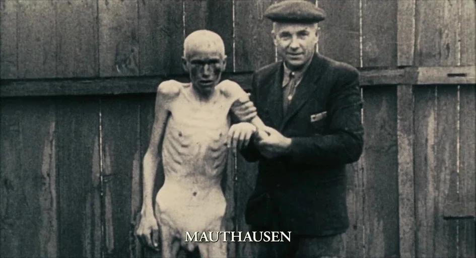The Last Days (1998) movie still: medium — An emaciated prisoner stands in front of a rustic wooden plank wall. A man in a suit grips the prisoner's…