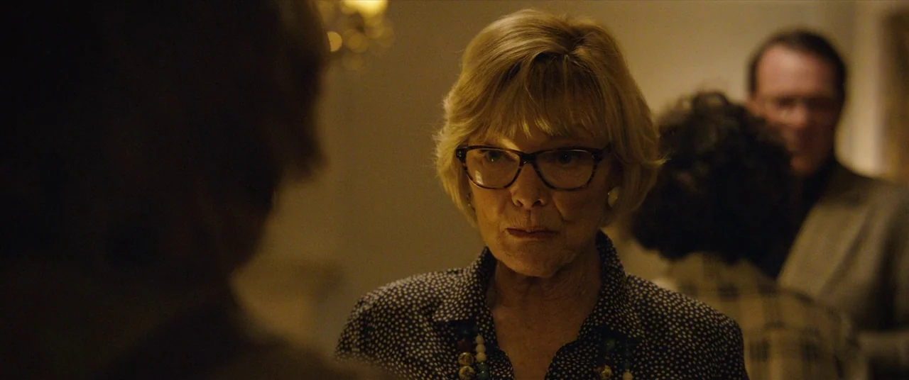 Can You Ever Forgive Me? (2018) movie still: closeup — An older woman stands in a dimly lit, crowded interior space with soft background blur. She…