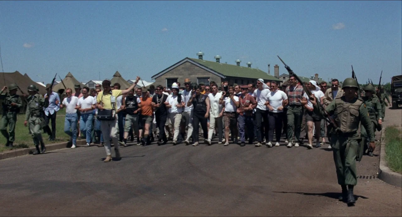 Medium Cool (1969) movie still: wide — A group of men in casual clothing walk down a road on a military base. Several armed soldiers in green…