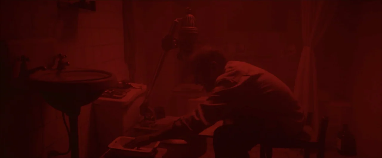 My Neighbor Adolf (2022) movie still: medium — A small bathroom converted into a darkroom with a sink and photo equipment. A man sits on a stool and…