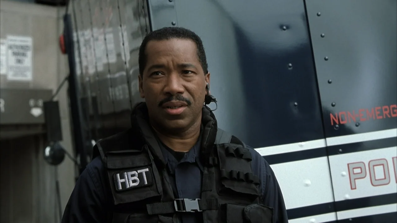 John Q (2002) movie still: medium — An exterior police scene features a man in tactical gear standing before a van. He wears a dark uniform and a…