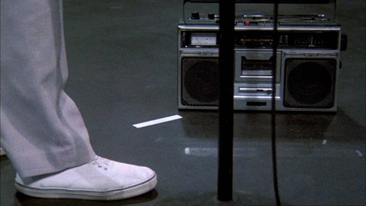 Stop Making Sense (1984) movie still: medium — A spacious room with a polished, dark gray floor featuring a lone boombox. A person wearing white…