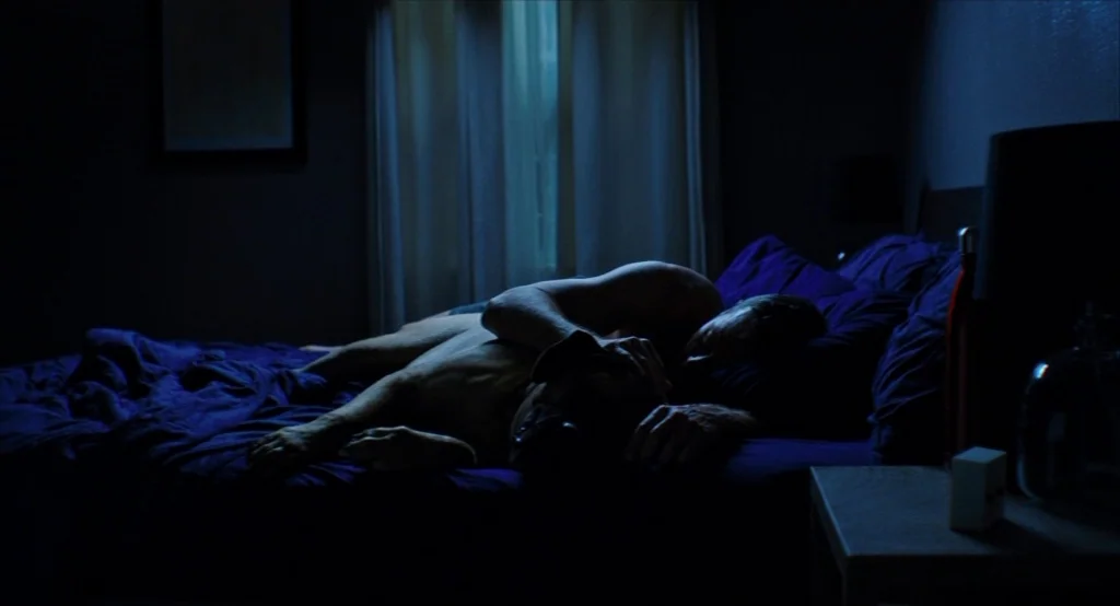 Results (2015) movie still: medium — A dark bedroom with blue sheets and a bedside table with a small light. A man lies in bed with his arm around a…