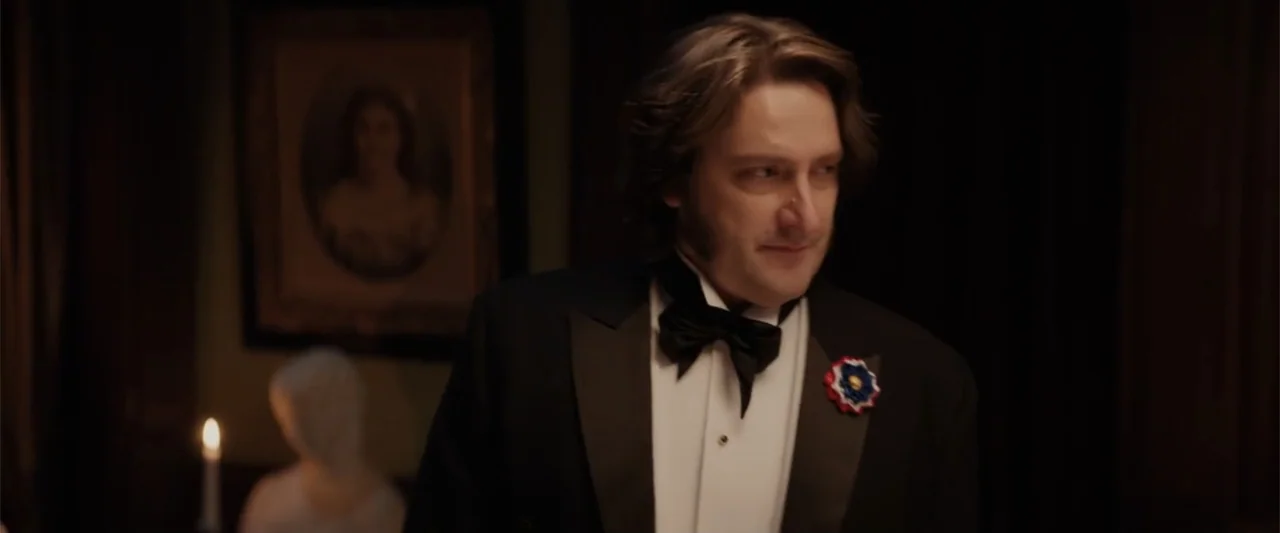 I Heard the Bells (2022) movie still: medium — A dark parlor room features a framed portrait on the wall and a lit candle. A man in formal evening…