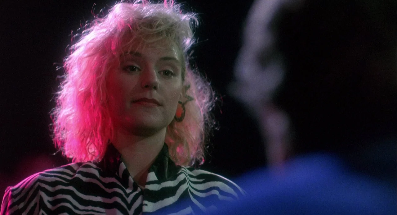 Vamp (1986) movie still: closeup — A dimly lit nightclub with a dark, indistinct background. A young woman with blonde hair stands in the…