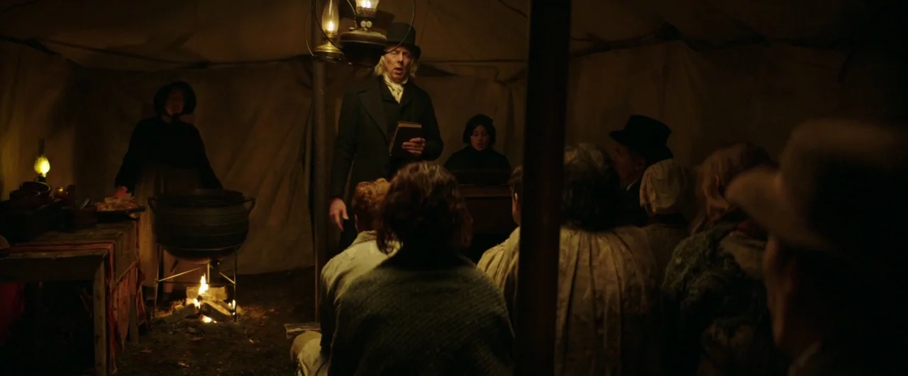 Black ’47 (2018) movie still: wide — A dimly lit canvas tent contains a group of people listening to a speaker. An elderly man in a top hat holds a…