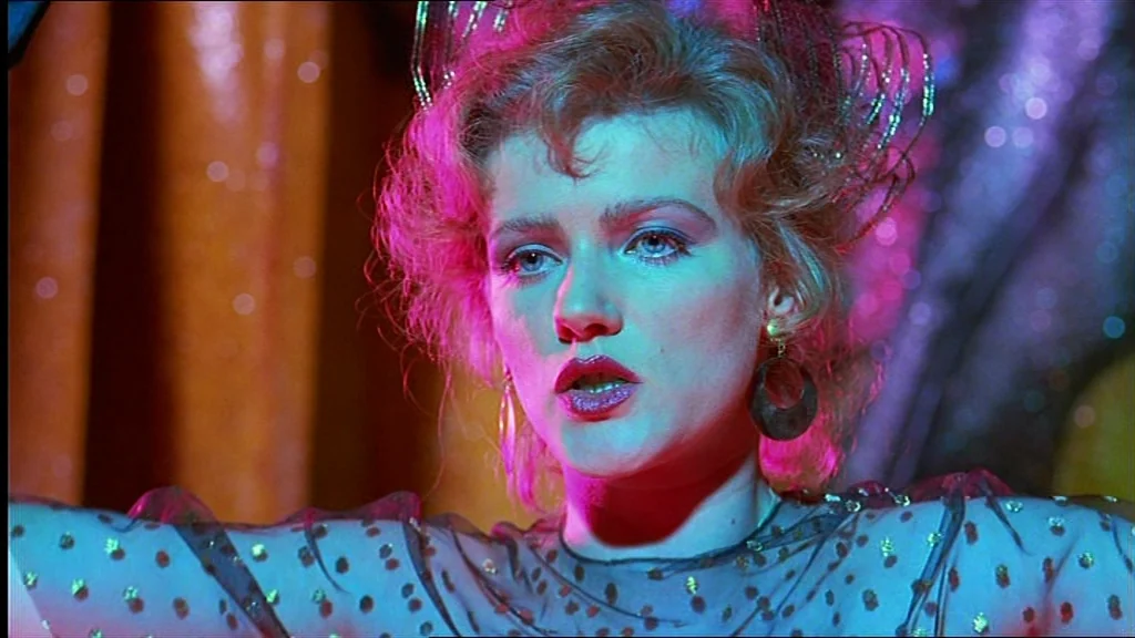 Lola (1981) movie still: closeup — A dimly lit interior space with reflective surfaces and blurred orange and purple lights. A woman faces the…