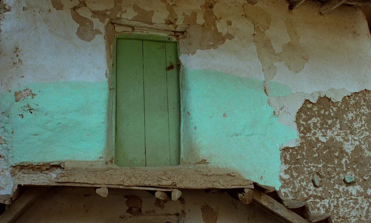 Through The Olive Trees (1994) movie still: medium — An aged adobe wall features peeling plaster and a single weathered green wooden door. The wall…