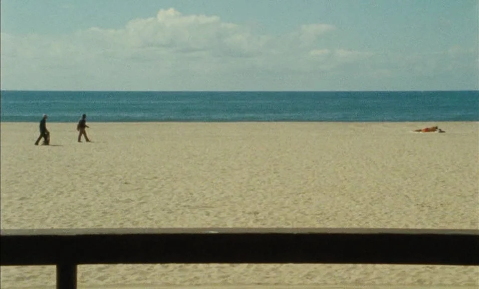 Documenteur (1981) movie still: wide — A wide sandy beach stretches toward the calm blue ocean under a partly cloudy sky. Two people walk across the…