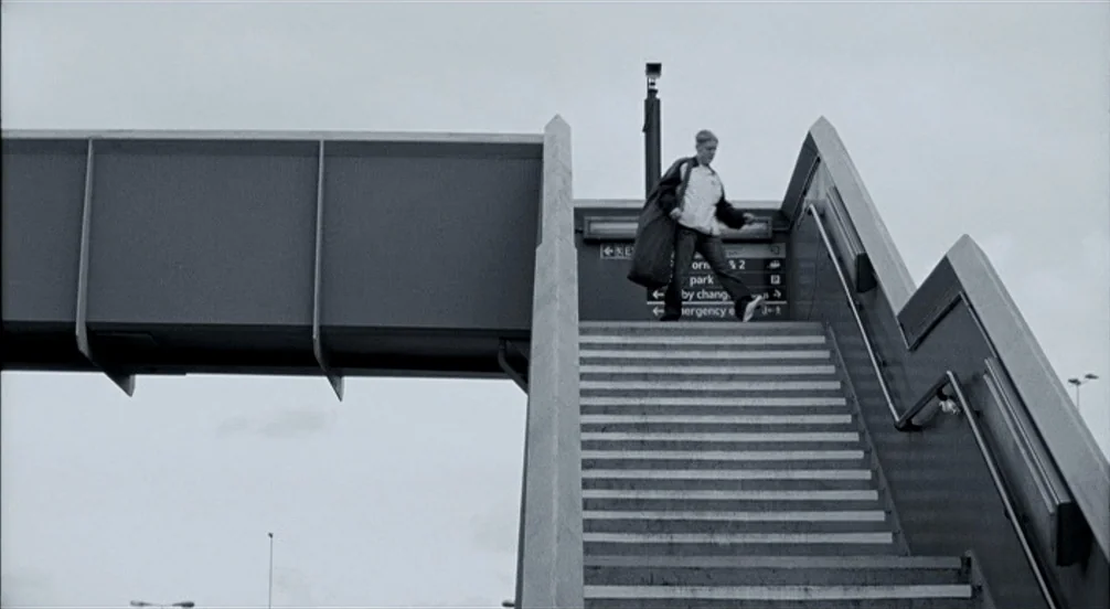 Somers Town (2008) movie still: wide — A modern concrete footbridge extends over a rail station area with navigational signage. A young man walks…