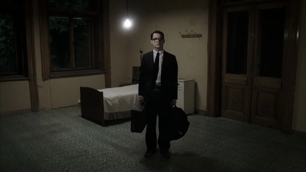Everything Is Illuminated (2005) movie still: fullbody — A sparsely furnished room contains a single bed and a wooden door. A young man in a suit…