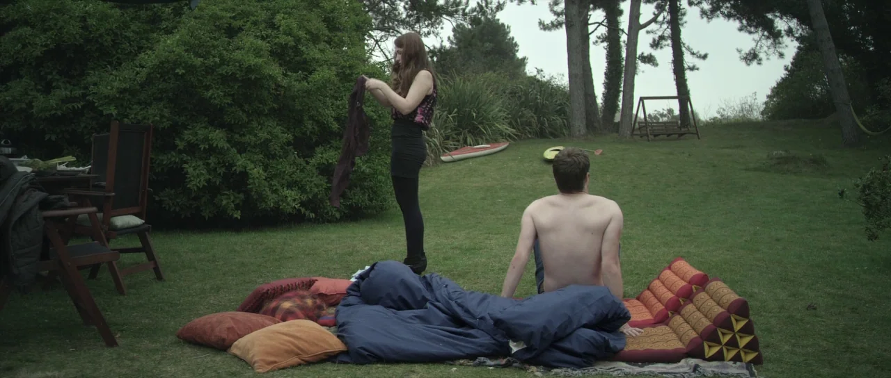 What Richard Did (2012) movie still: wide — A grassy backyard features a bedding setup on the lawn near dense bushes. A woman stands holding a dark…