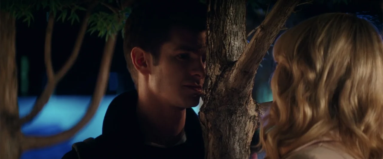 The Amazing Spider-Man 2 (2014) movie still: medium — A nighttime outdoor setting with thick tree trunks and a dark background. Two young people…