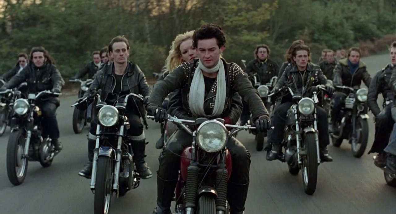 Quadrophenia (1979) movie still: wide — A group of motorcycle riders travels along an asphalt road lined by dense trees. The lead rider steers a red…