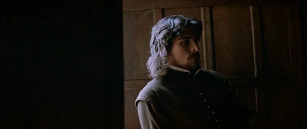 The New World (2005) movie still: medium — A man stands in a dimly lit, wood-paneled room near a dark doorway. He is angled away from the camera,…