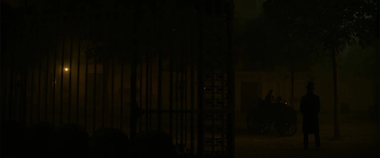 Eiffel (2021) movie still: wide — A dark city street at night is partially blocked by a large iron gate. A man in a top hat stands near a…