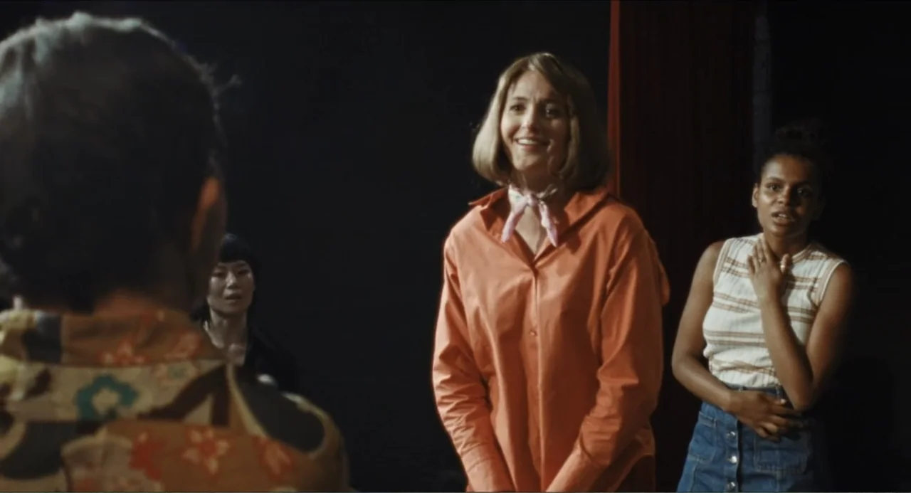 A Different Man (2024) movie still: medium — A dark, sparsely furnished room with a red curtain partially visible on the right. Three women stand…