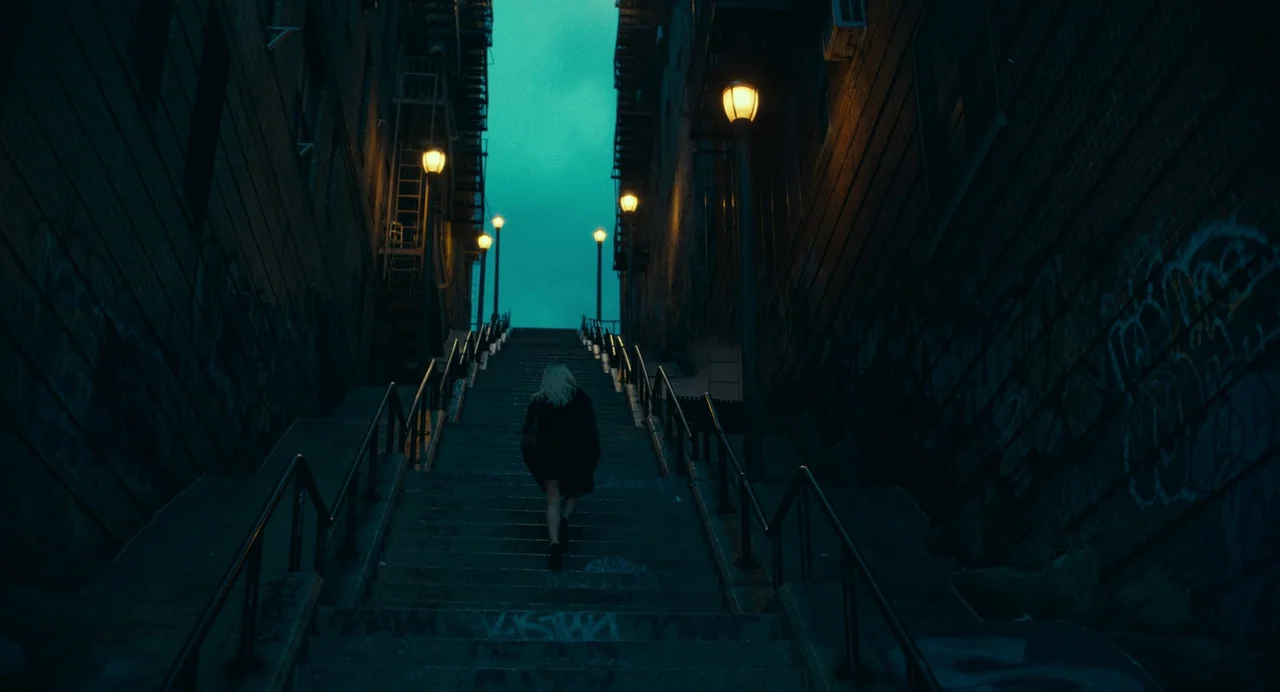 Joker: Folie à Deux (2024) movie still: wide — An urban outdoor staircase flanked by tall brick walls covered in graffiti at night. A young woman…